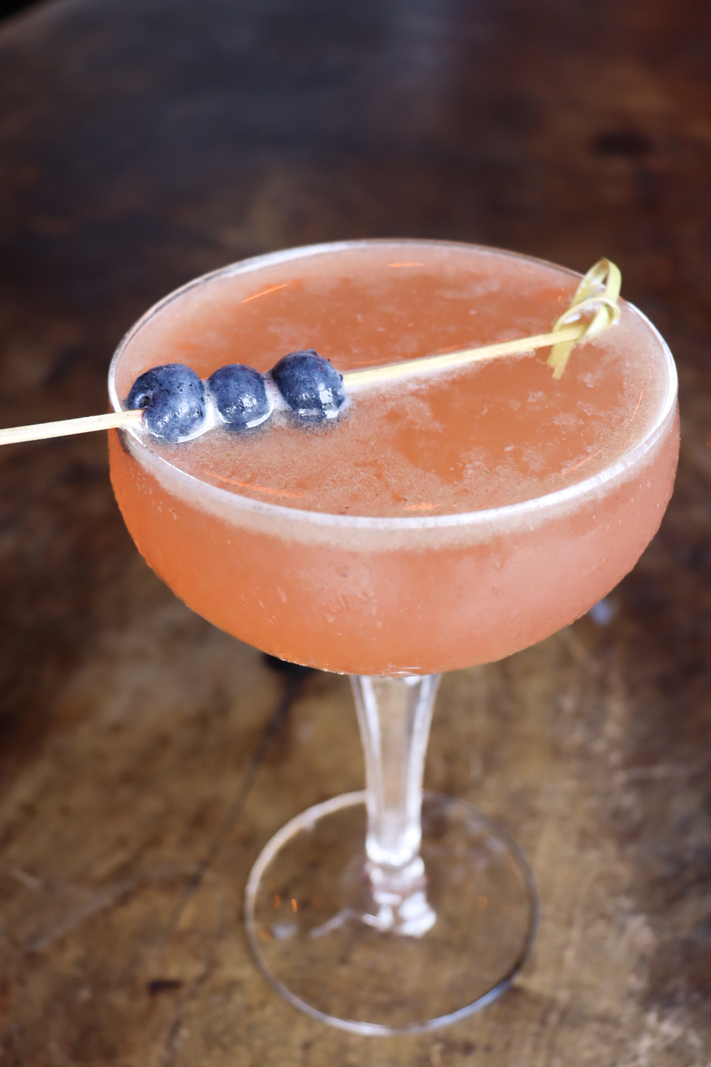 Blueberry Garnish Cocktail Photography – Tampa Restaurant Photographer Orange cocktail with blueberry garnish in coupe glass on wooden table