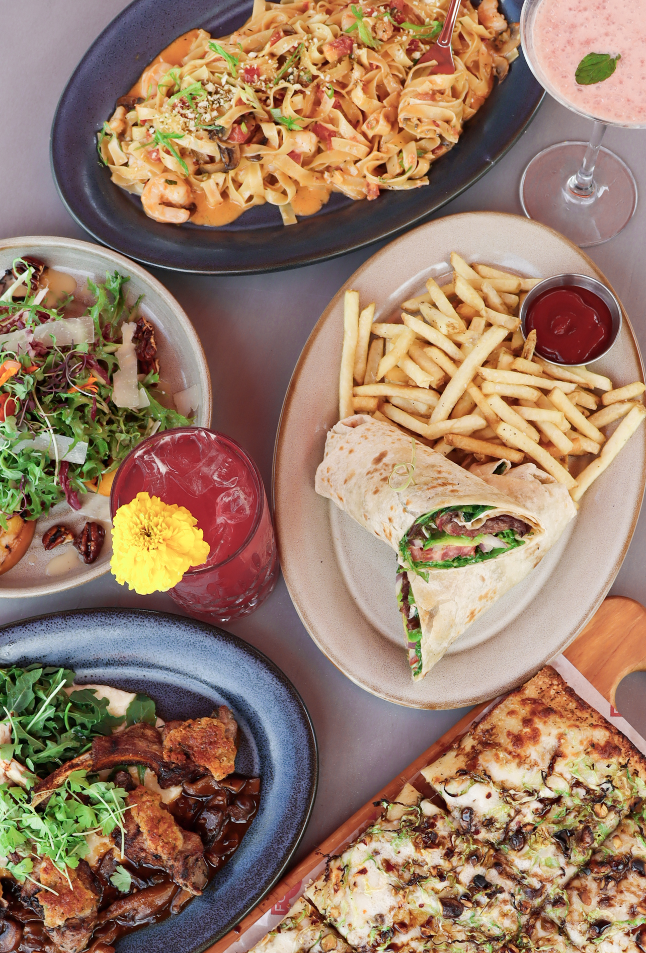 Overhead view of assorted restaurant dishes including pasta, wrap with fries, flatbread, cocktails, and tacos