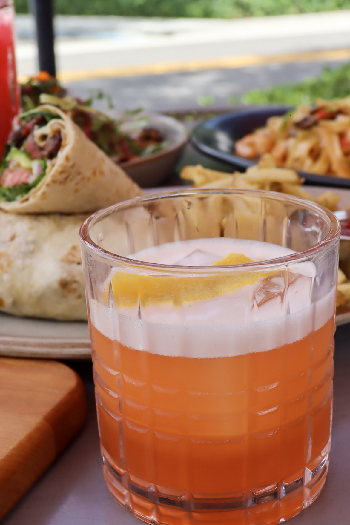 Cocktail & Food Pairing Photography – Tampa Restaurant Content Orange cocktail with lemon garnish in textured glass with wraps and pasta in background